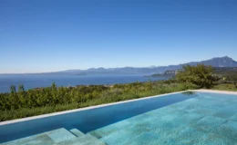 Stunning view of a modern infinity pool overlooking a serene lake and mountainous landscape under a clear blue sky, surrounded by lush greenery. Ideal for luxury vacation retreats and scenic getaways.