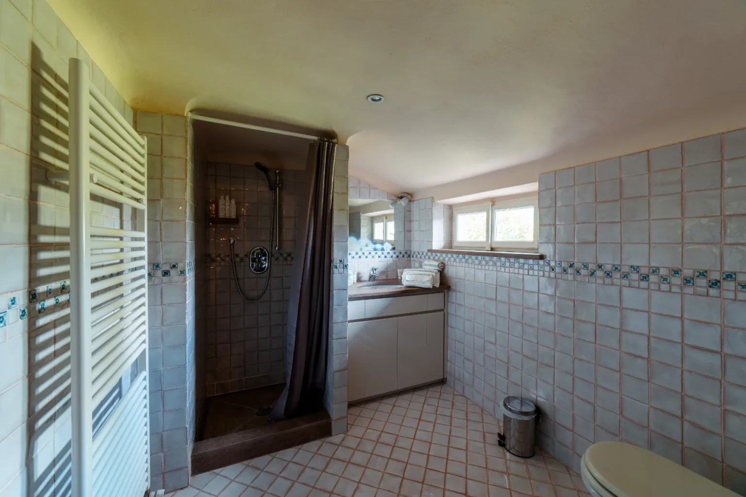 Spacious bathroom featuring a shower area with a gray curtain, modern fixtures, and tiled walls. Includes a wooden countertop with neatly arranged towels, a sink, and a trash can, complemented by natural light from windows. Ideal for relaxation and comfort. | Only on SopranoVillas