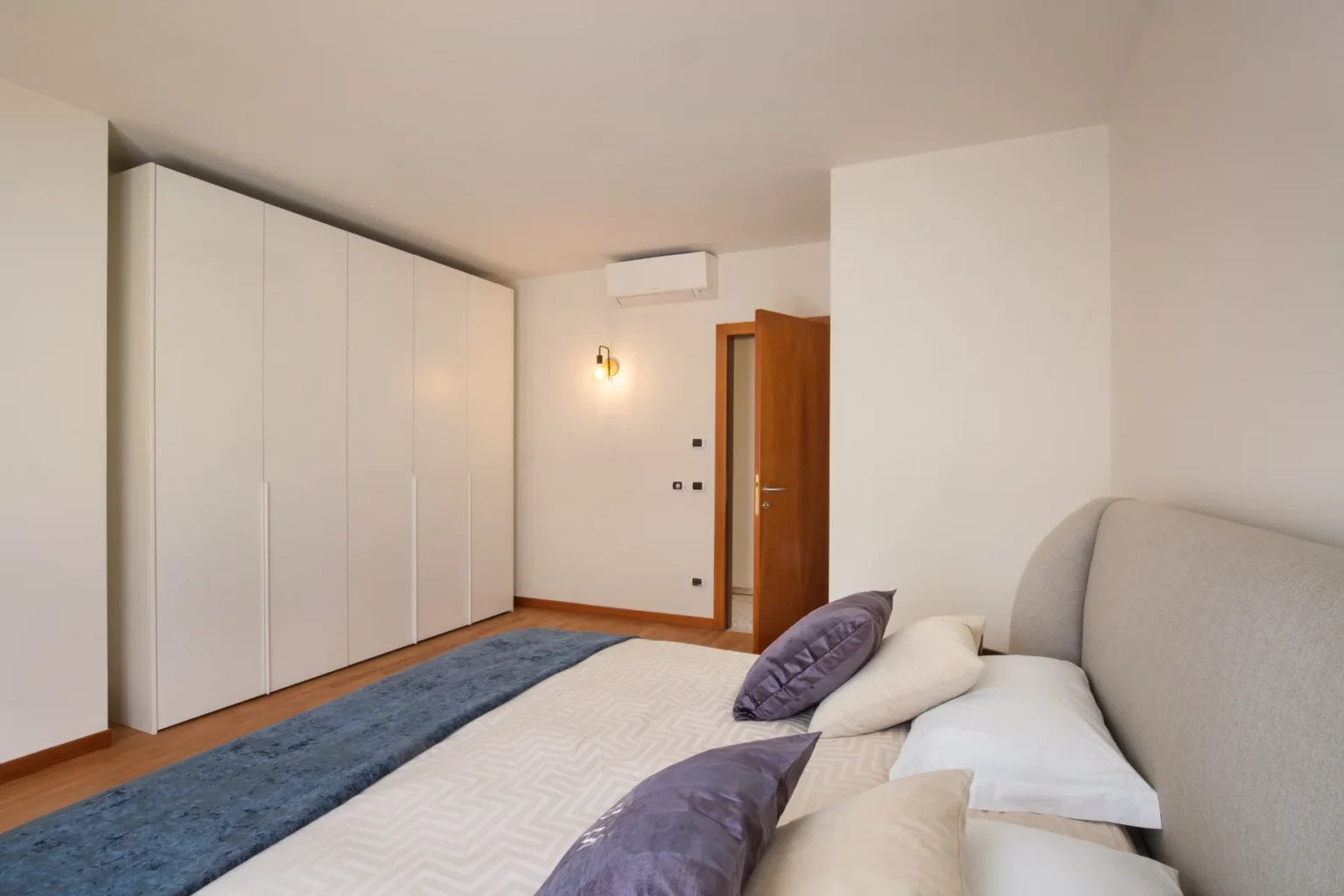 Modern bedroom interior featuring a neatly made bed with decorative pillows, a light-colored wall, and a spacious white wardrobe. Natural light illuminates the room, highlighting the wooden floor and stylish door. | Only on SopranoVillas