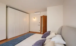 Modern bedroom interior featuring a neatly made bed with decorative pillows, a light-colored wall, and a spacious white wardrobe. Natural light illuminates the room, highlighting the wooden floor and stylish door.