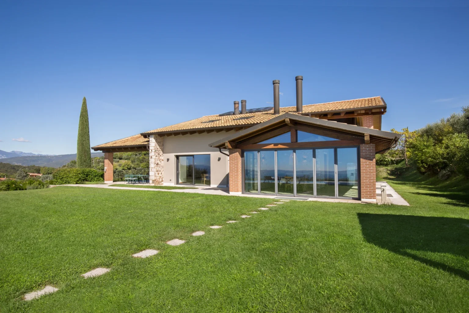 Modern hillside villa featuring large glass windows, a sloped roof, and a spacious lawn with stone pathway, surrounded by lush greenery and mountain views. Perfect for luxury living and outdoor relaxation. | Only on SopranoVillas