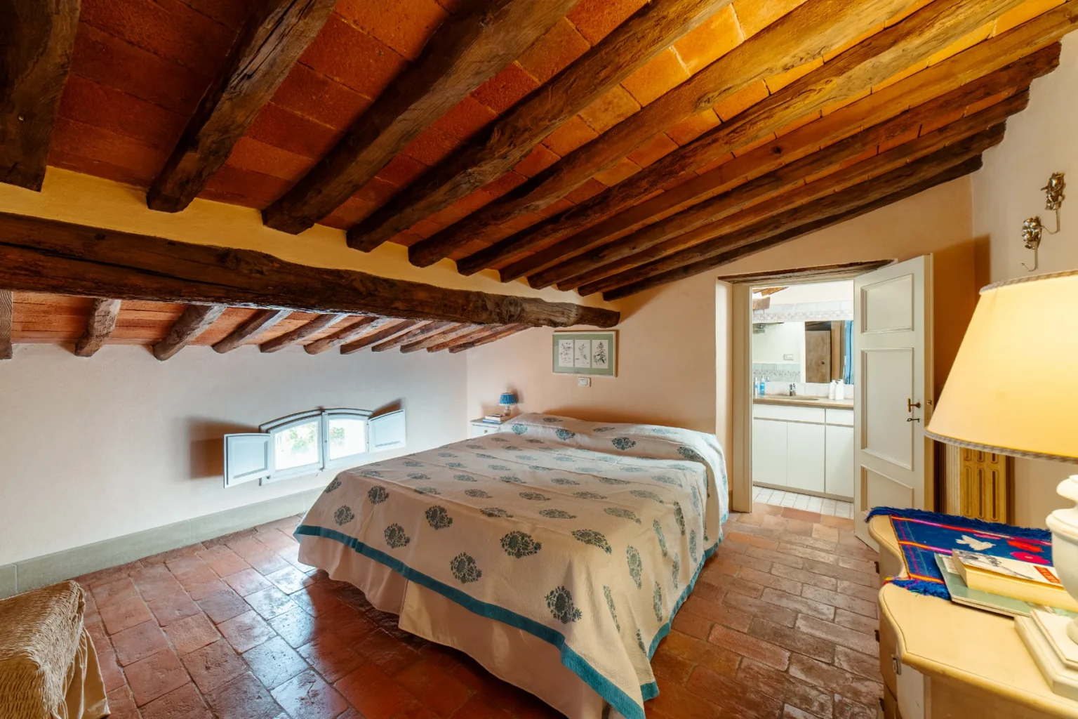 Cozy bedroom with a rustic design featuring wooden beams and terracotta tiles, a large bed with a floral-patterned quilt, and a window offering natural light. The room includes a bedside table and a doorway leading to a bathroom area. Ideal for a relaxing getaway. | Only on SopranoVillas