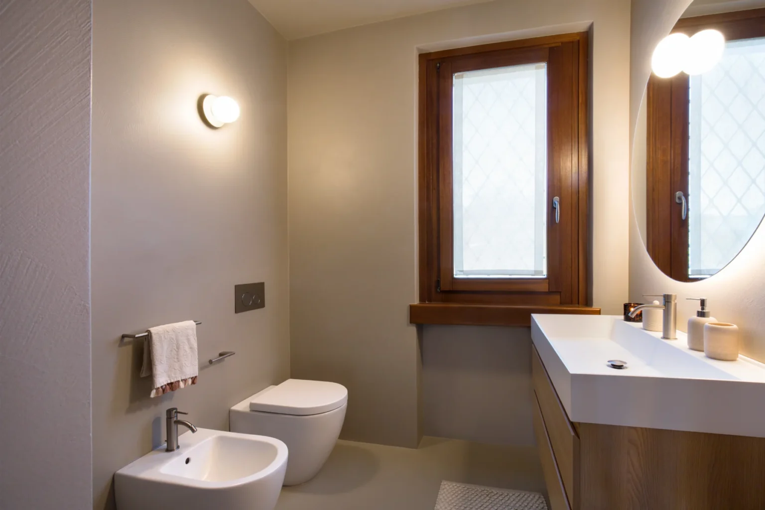 Modern bathroom featuring a minimalist design with a wall-mounted toilet, bidet, and a sleek vanity with a round mirror. Natural light streams in through a window with wooden frames, complemented by neutral wall colors and contemporary fixtures. | Only on SopranoVillas