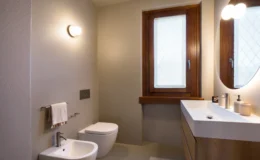 Modern bathroom featuring a minimalist design with a wall-mounted toilet, bidet, and a sleek vanity with a round mirror. Natural light streams in through a window with wooden frames, complemented by neutral wall colors and contemporary fixtures.
