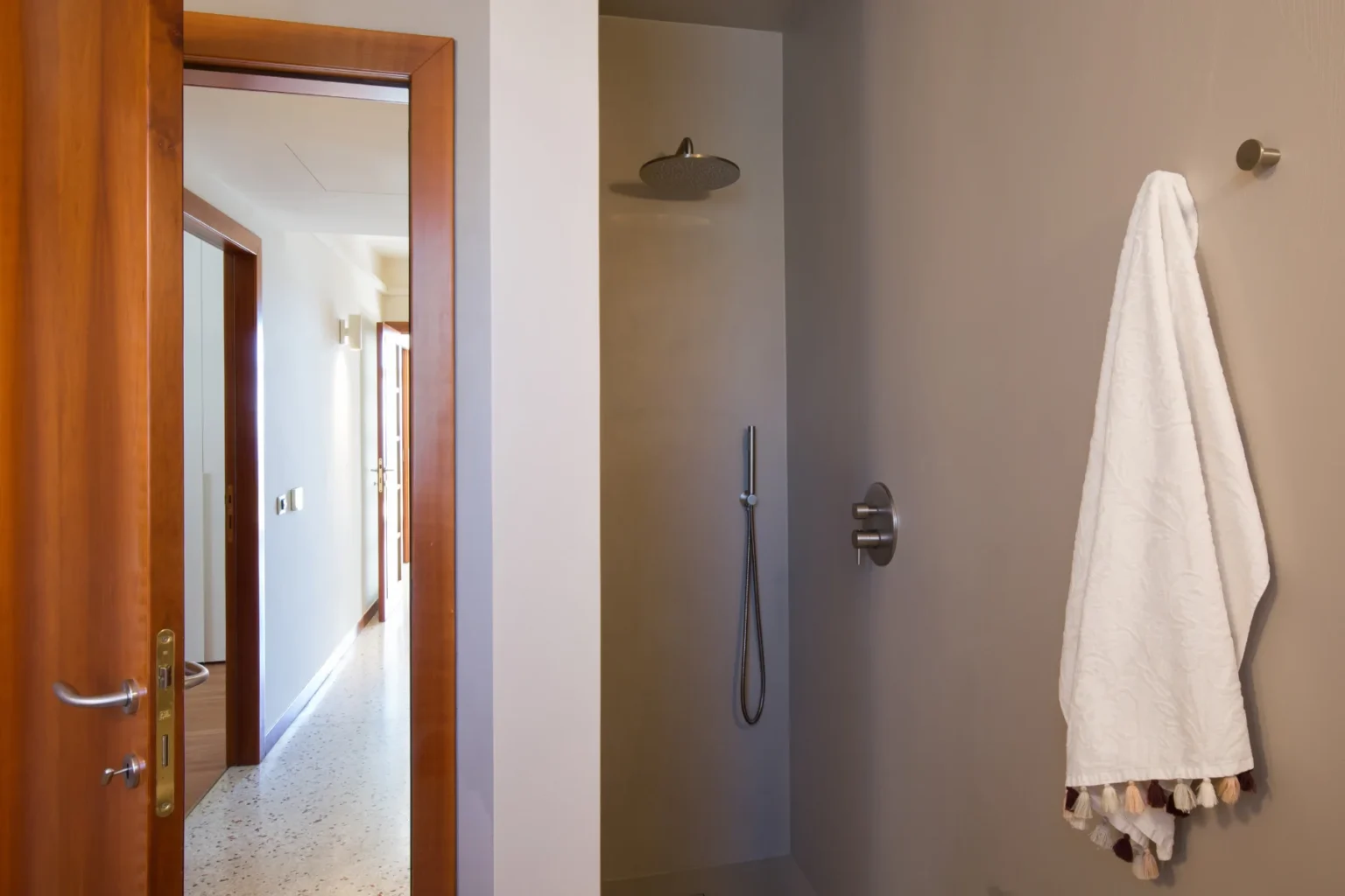 Modern bathroom shower area featuring a sleek, minimalist design with a rain showerhead, handheld shower, and a plush white towel hanging on the wall. A wooden door leads to a well-lit hallway, showcasing a clean and elegant interior. | Only on SopranoVillas