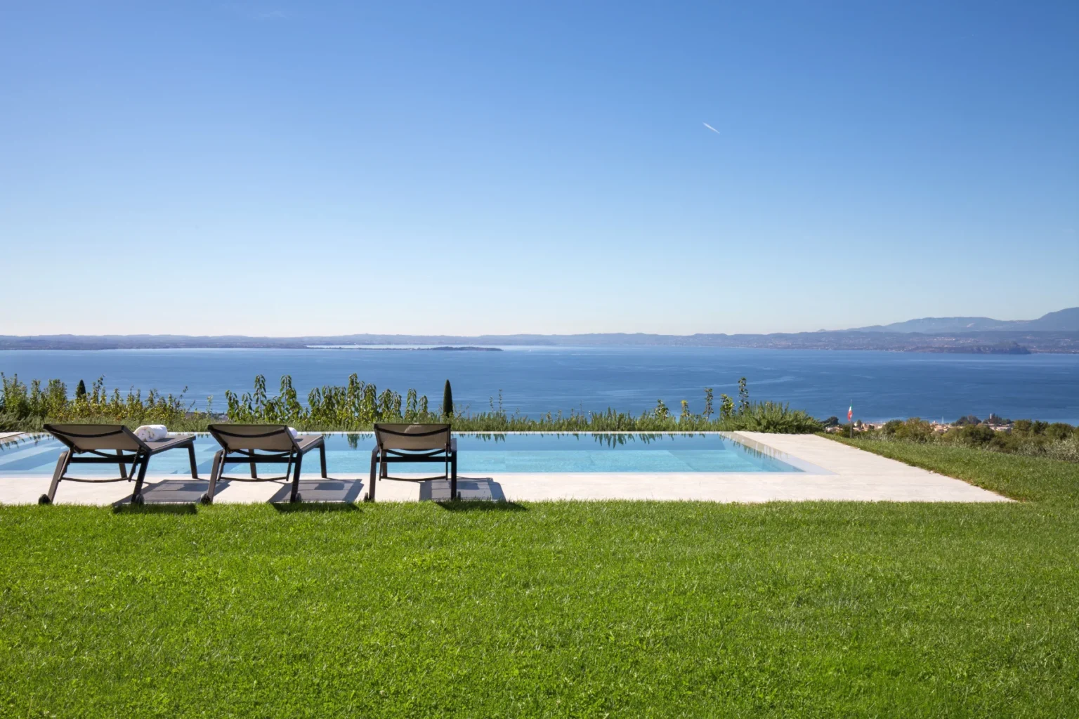 Scenic view of a luxurious infinity pool overlooking a calm blue sea, with three lounge chairs on a manicured green lawn under a clear blue sky. Ideal for relaxation and outdoor enjoyment. | Only on SopranoVillas