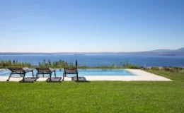 Scenic view of a luxurious infinity pool overlooking a calm blue sea, with three lounge chairs on a manicured green lawn under a clear blue sky. Ideal for relaxation and outdoor enjoyment.
