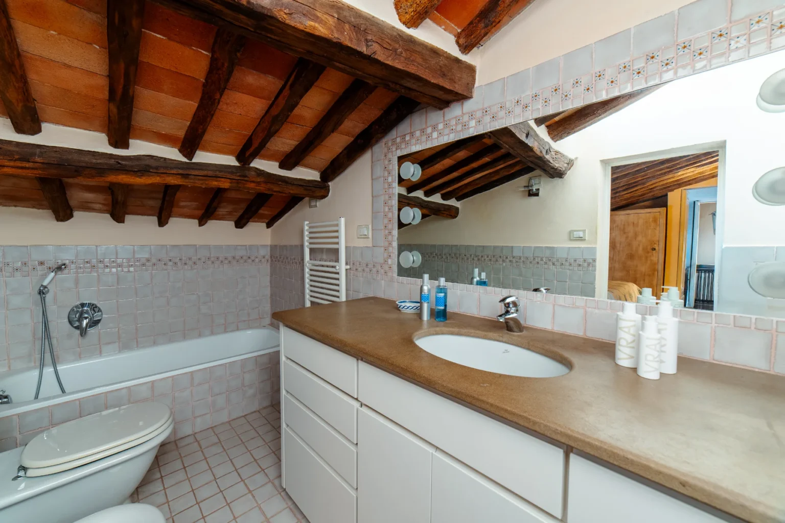 Cozy bathroom with wooden beams, featuring a bathtub, modern sink, and stylish vanity. The walls are adorned with light-colored tiles, and there are various toiletries neatly arranged on the countertop. Natural light illuminates the space, enhancing its inviting atmosphere. | Only on SopranoVillas