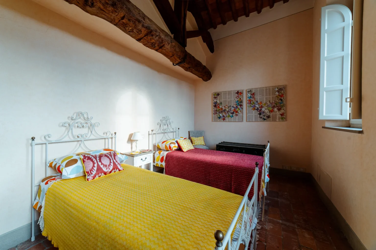 Bright and cozy bedroom featuring two twin beds with colorful blankets, a bedside table with a lamp, and decorative pillows. The room showcases rustic wooden beams and large windows, creating a warm and inviting atmosphere. Art pieces adorn the walls, adding character to the space. Ideal for family or friends traveling together. | Only on SopranoVillas