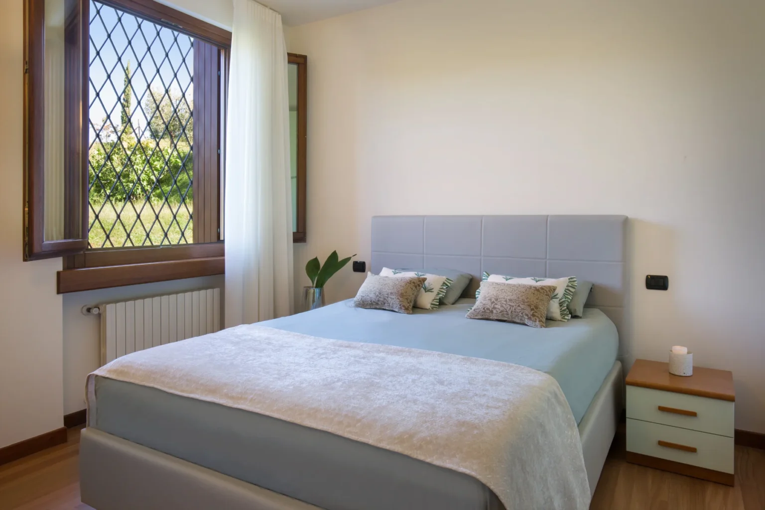 Modern bedroom featuring a comfortable bed with light blue bedding and decorative pillows, a stylish headboard, and a wooden nightstand. Natural light streams in through a large window with diamond-patterned panes, highlighting the serene outdoor view and a decorative plant nearby. | Only on SopranoVillas