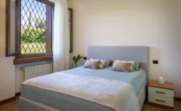 Modern bedroom featuring a comfortable bed with light blue bedding and decorative pillows, a stylish headboard, and a wooden nightstand. Natural light streams in through a large window with diamond-patterned panes, highlighting the serene outdoor view and a decorative plant nearby.