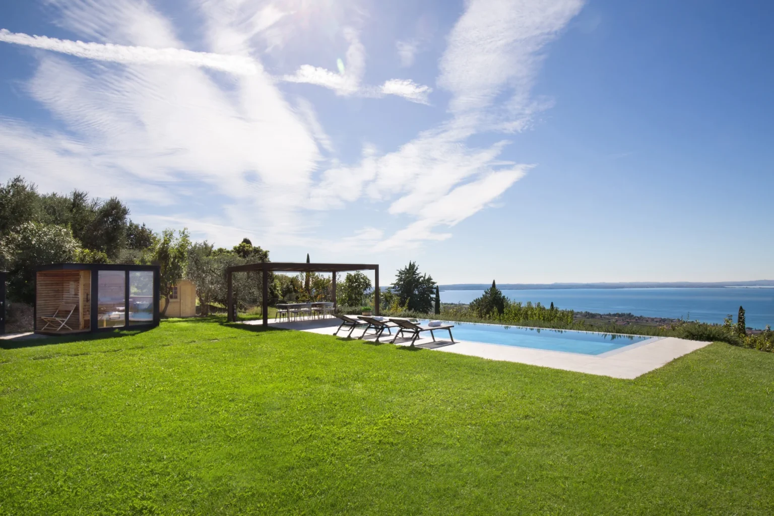 Scenic view of a modern outdoor space featuring a swimming pool, lounge chairs, and a pergola surrounded by lush green grass and trees, overlooking a tranquil sea under a clear blue sky. | Only on SopranoVillas