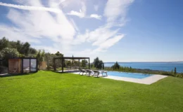 Scenic view of a modern outdoor space featuring a swimming pool, lounge chairs, and a pergola surrounded by lush green grass and trees, overlooking a tranquil sea under a clear blue sky.