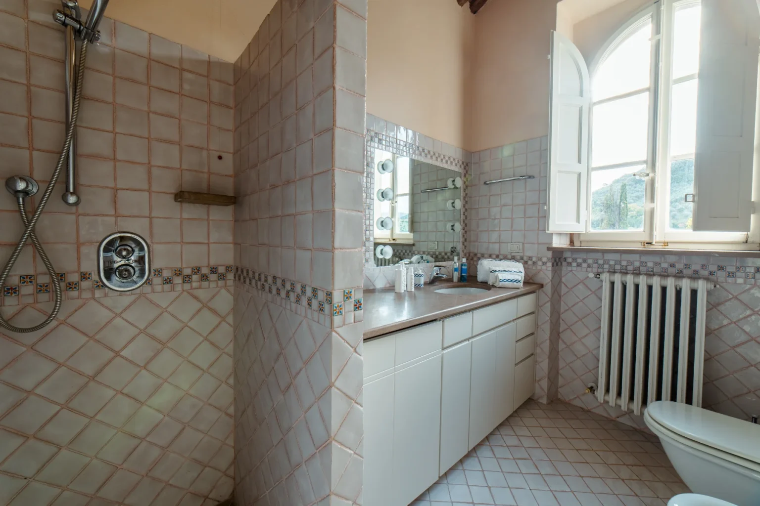 Bright and spacious bathroom featuring a modern shower with tiled walls, a large vanity with a mirror, and natural light streaming in through an open window. The decor includes pastel-colored tiles and essential toiletries arranged neatly on the countertop. | Only on SopranoVillas