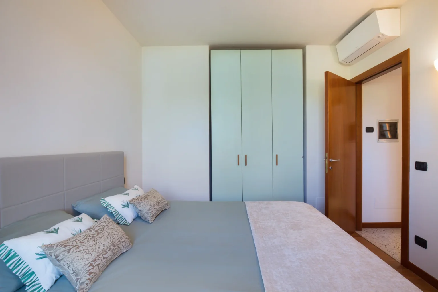 Modern bedroom featuring a comfortable bed with decorative pillows, a light blue wardrobe, and a wooden door leading to another room. The space is bright and airy, with an air conditioning unit mounted on the wall. | Only on SopranoVillas