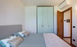 Modern bedroom featuring a comfortable bed with decorative pillows, a light blue wardrobe, and a wooden door leading to another room. The space is bright and airy, with an air conditioning unit mounted on the wall.