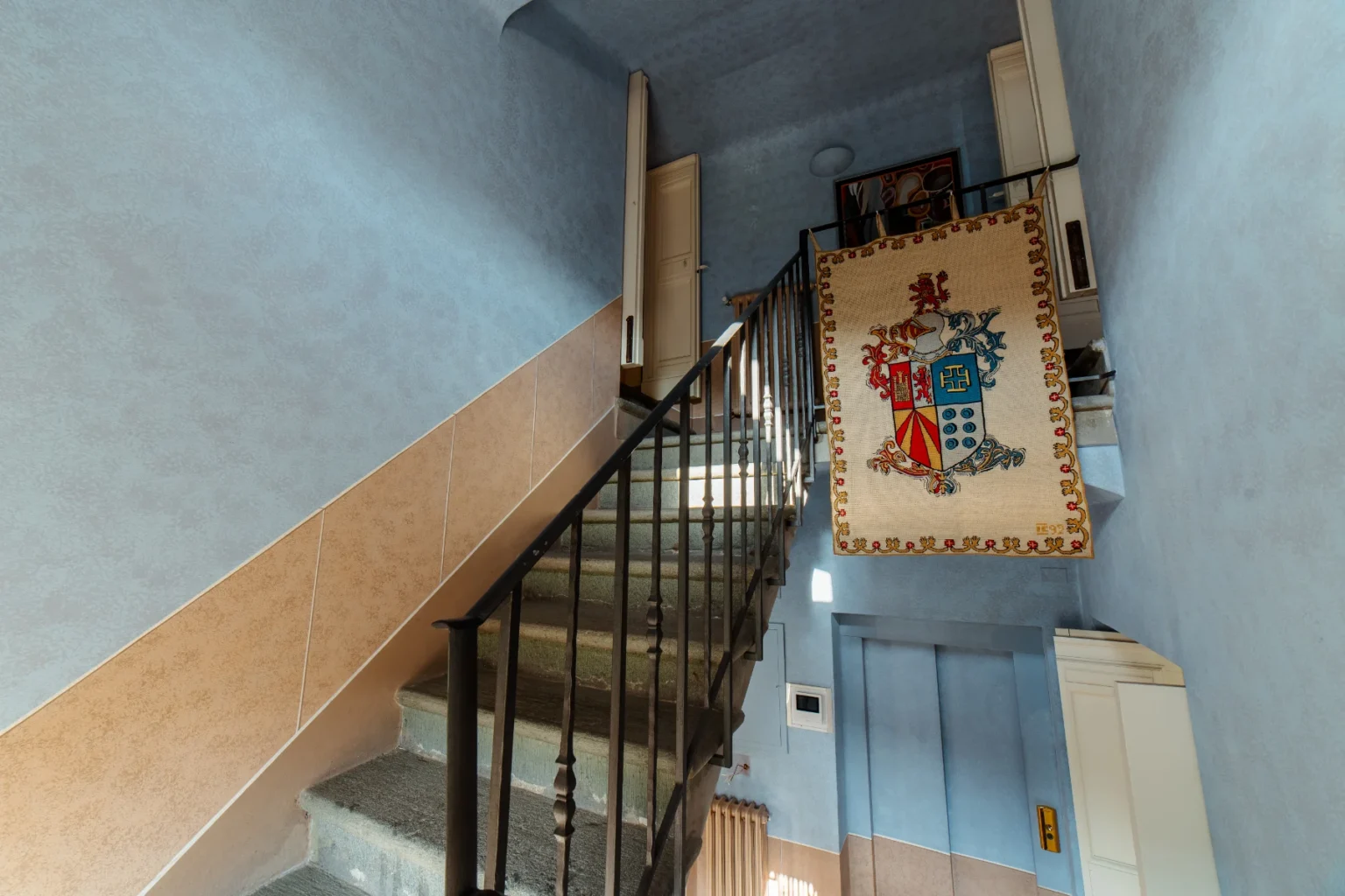 Staircase with blue and beige walls, featuring a decorative tapestry with a coat of arms hanging from the railing, illuminated by natural light. | Only on SopranoVillas