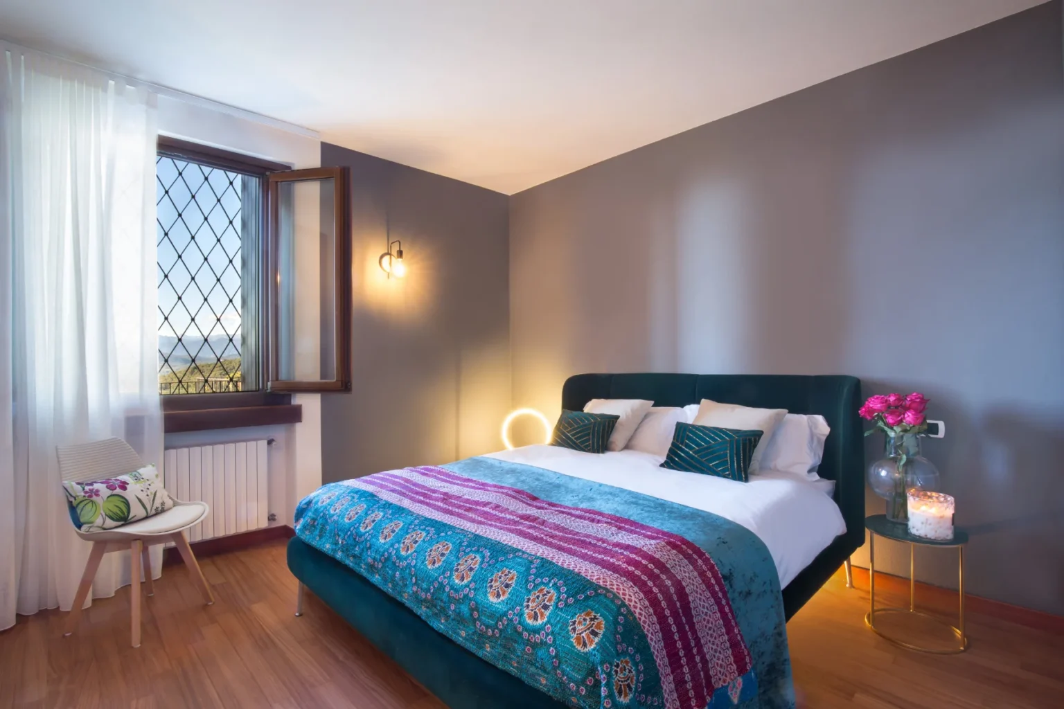 Cozy bedroom featuring a green upholstered bed with decorative pillows, a colorful throw blanket, and a stylish bedside table with a vase of pink roses and a candle. Natural light streams in through a large window adorned with sheer curtains, highlighting the warm wooden flooring and modern decor elements. | Only on SopranoVillas