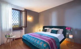 Cozy bedroom featuring a green upholstered bed with decorative pillows, a colorful throw blanket, and a stylish bedside table with a vase of pink roses and a candle. Natural light streams in through a large window adorned with sheer curtains, highlighting the warm wooden flooring and modern decor elements.