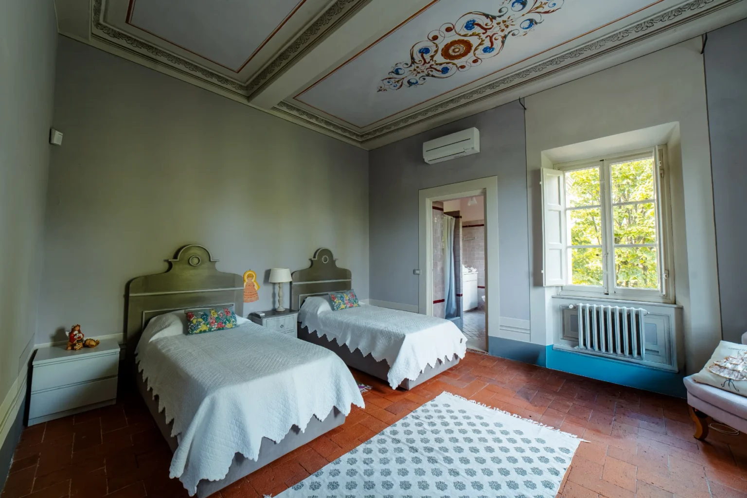 Spacious bedroom featuring two twin beds with decorative headboards, a cozy lamp, and colorful accents. The room has a large window with natural light, showcasing a view of greenery outside. Elegant ceiling design and warm terracotta flooring create a welcoming atmosphere. | Only on SopranoVillas