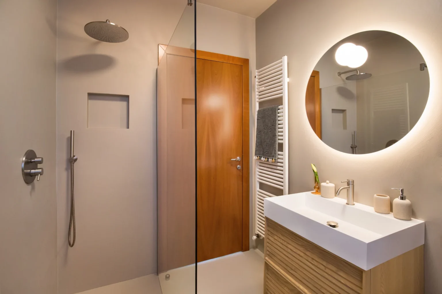 Modern bathroom featuring a glass shower, sleek wooden door, and minimalist vanity with a round mirror. The space includes a towel radiator and elegant fixtures, showcasing a contemporary design aesthetic. | Only on SopranoVillas