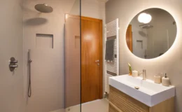 Modern bathroom featuring a glass shower, sleek wooden door, and minimalist vanity with a round mirror. The space includes a towel radiator and elegant fixtures, showcasing a contemporary design aesthetic.