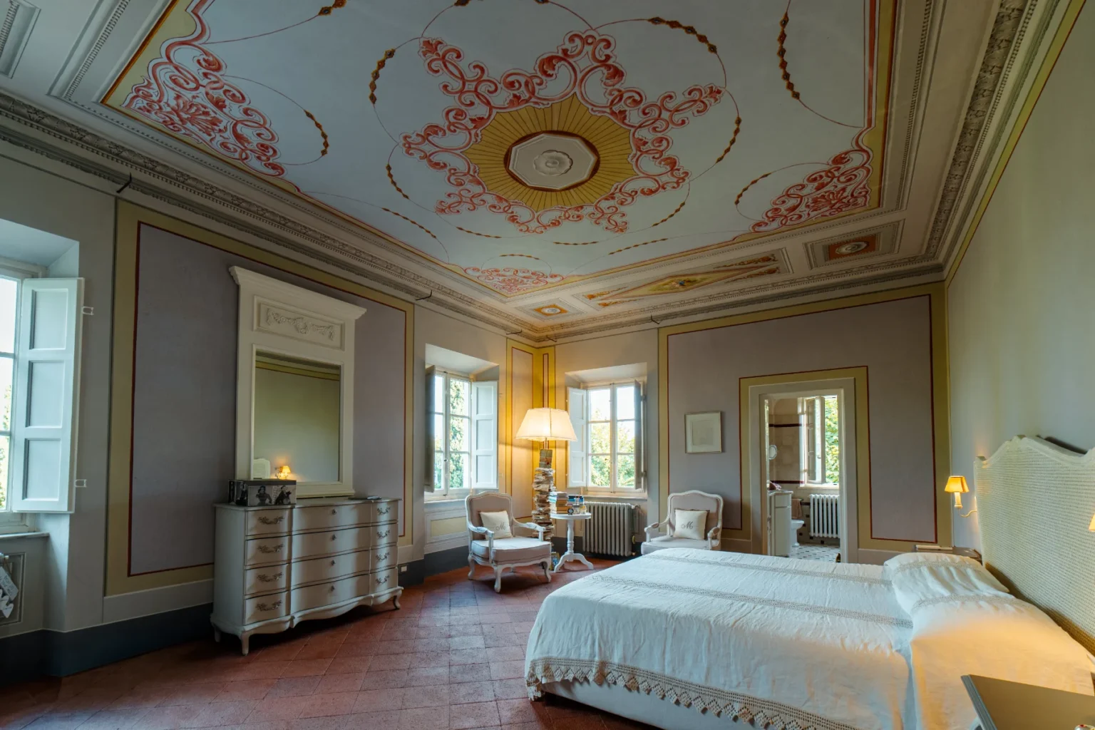 Elegant bedroom featuring a beautifully painted ceiling, classic furnishings, and natural light from large windows. The room includes a double bed with white linens, a vintage dresser with a mirror, and cozy seating areas, creating a serene and inviting atmosphere. | Only on SopranoVillas