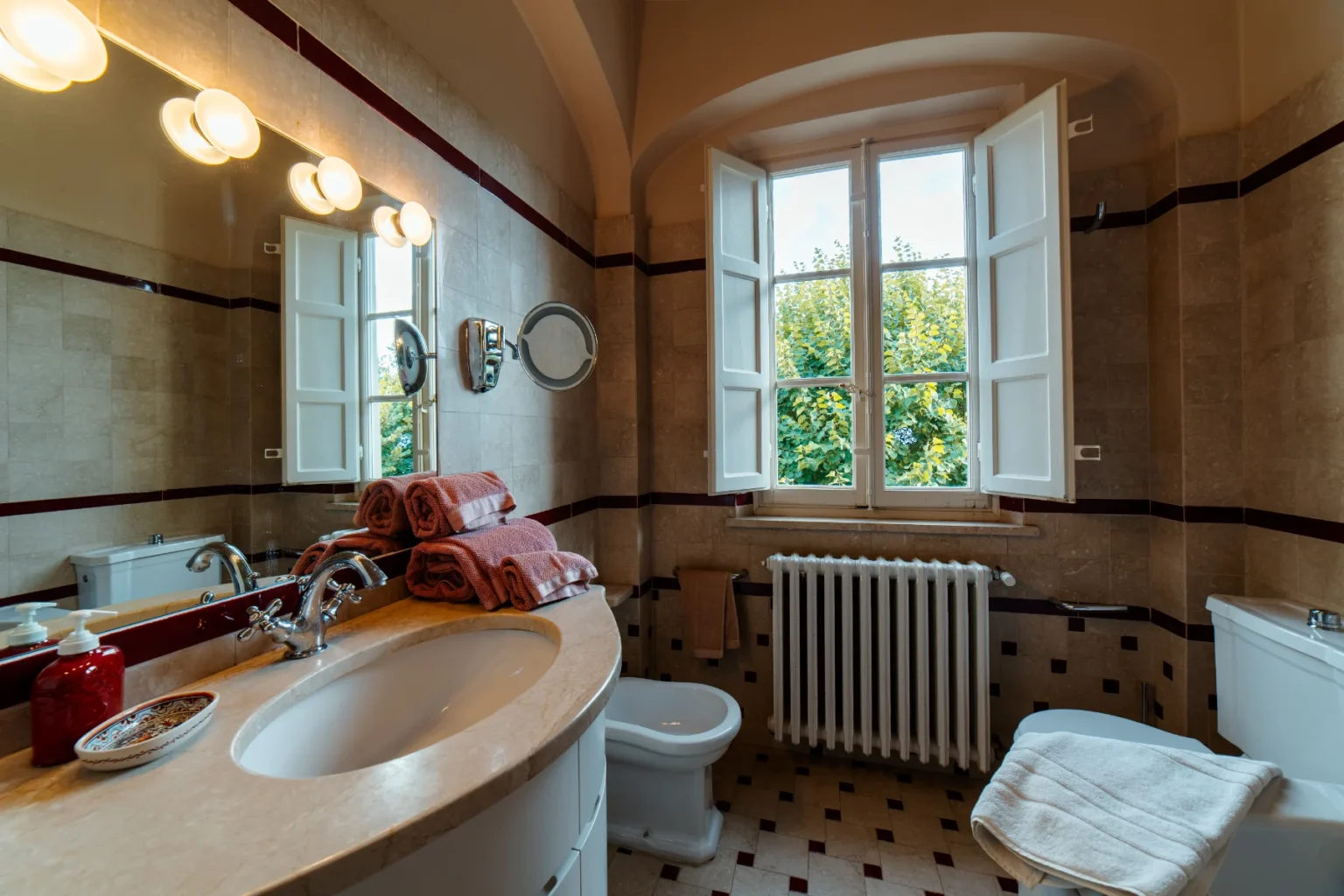 Elegant bathroom featuring a curved vanity with a sink, neatly stacked pink towels, a large mirror with lighting, and a window overlooking greenery. The decor includes beige tiles with maroon accents, a white toilet, and a radiator, creating a warm and inviting atmosphere. | Only on SopranoVillas