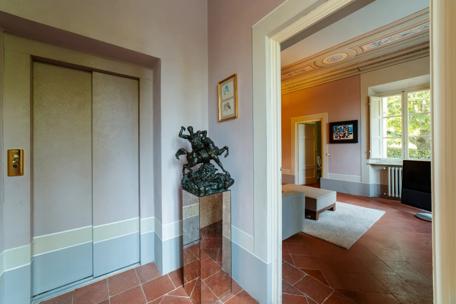 Interior view of a stylish hallway featuring an elevator, a bronze sculpture of a horseman on a pedestal, and a glimpse of a modern living area with large windows and elegant decor. | Only on SopranoVillas