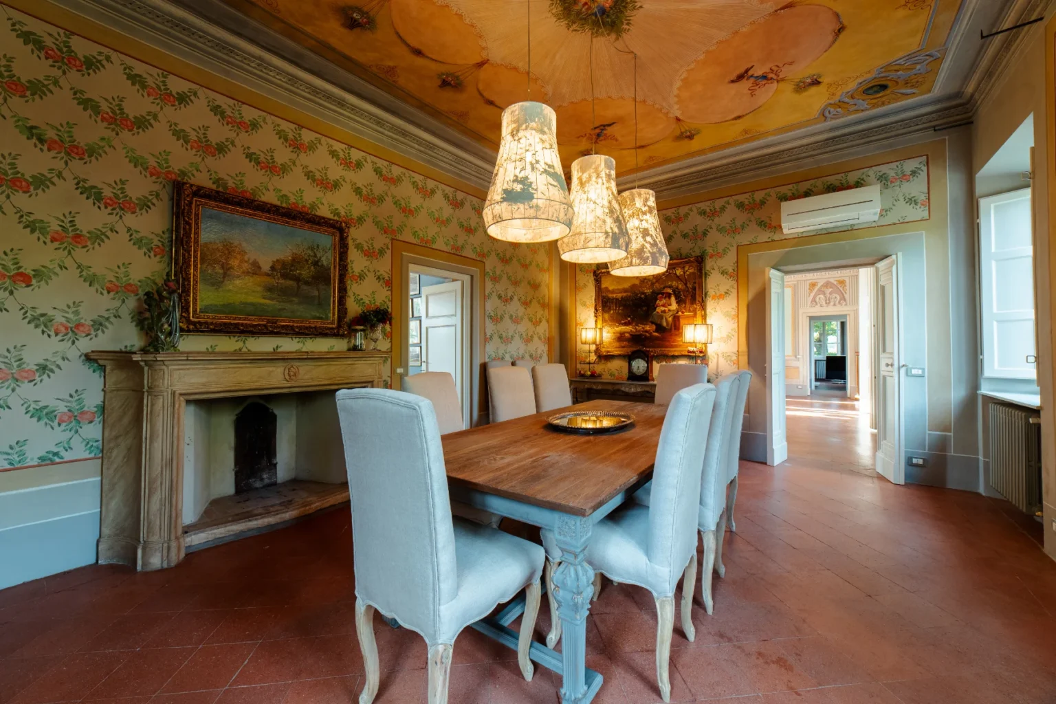Elegant dining room featuring a wooden table surrounded by plush chairs, adorned with stylish pendant lights. The walls are decorated with floral wallpaper, and a vintage painting hangs above a classic fireplace, creating a warm and inviting atmosphere. Natural light flows through the open doorway, enhancing the room's charm. | Only on SopranoVillas