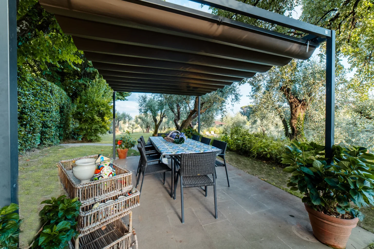 Outdoor dining area with a covered patio featuring a large table and chairs, surrounded by lush greenery and olive trees, ideal for alfresco meals. Decorative baskets and potted plants enhance the inviting atmosphere. | Only on SopranoVillas