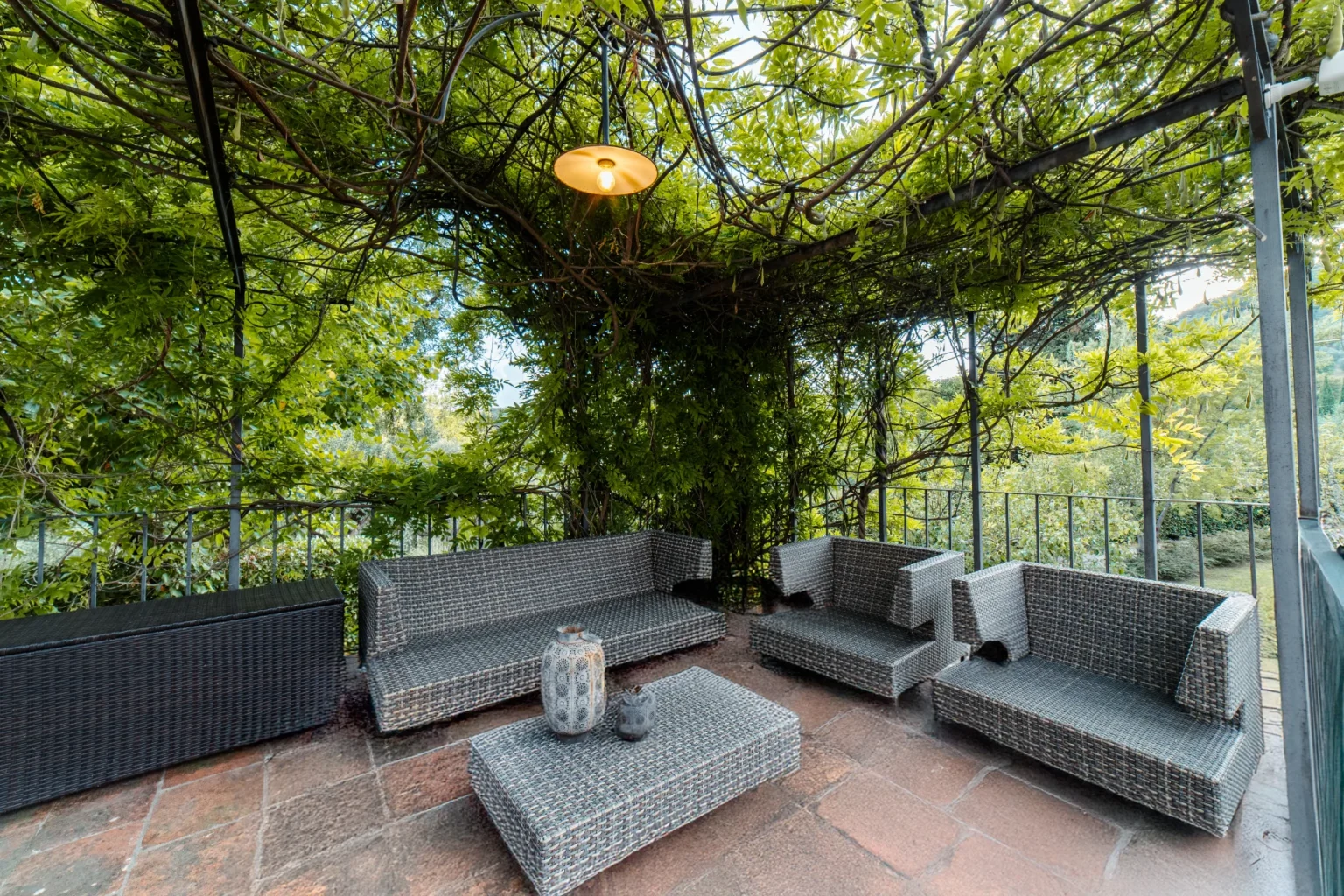 Cozy outdoor seating area featuring wicker furniture under a lush green canopy, perfect for relaxation and entertaining. The space includes a coffee table and decorative vases, surrounded by vibrant foliage and natural light. | Only on SopranoVillas