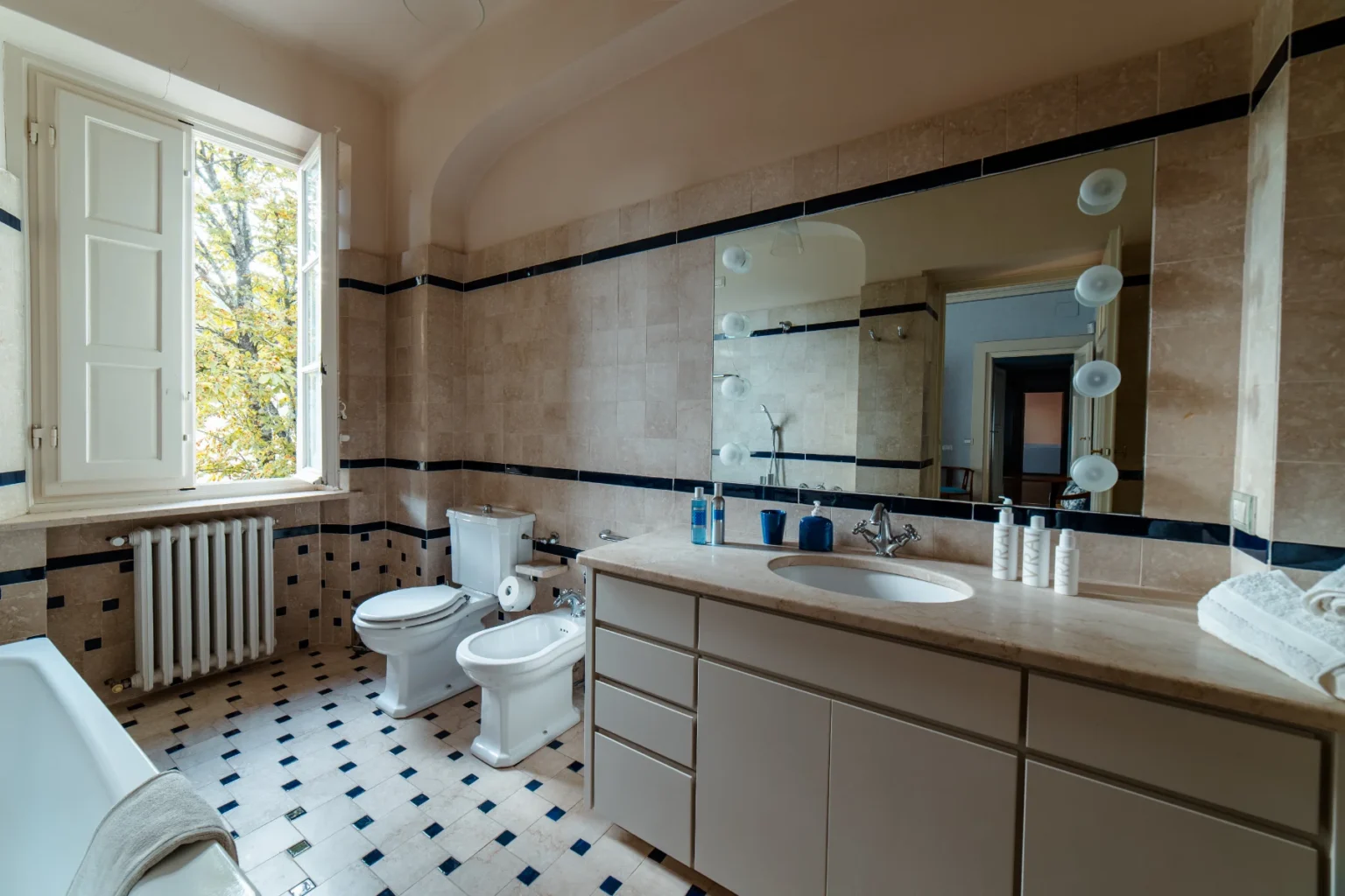 Bright and elegant bathroom featuring a large window with a view of greenery, beige tiled walls with navy accents, a modern vanity with a round sink, a bathtub, and two toilets. The space is well-lit with a mirror and stylish lighting, creating a serene atmosphere. | Only on SopranoVillas