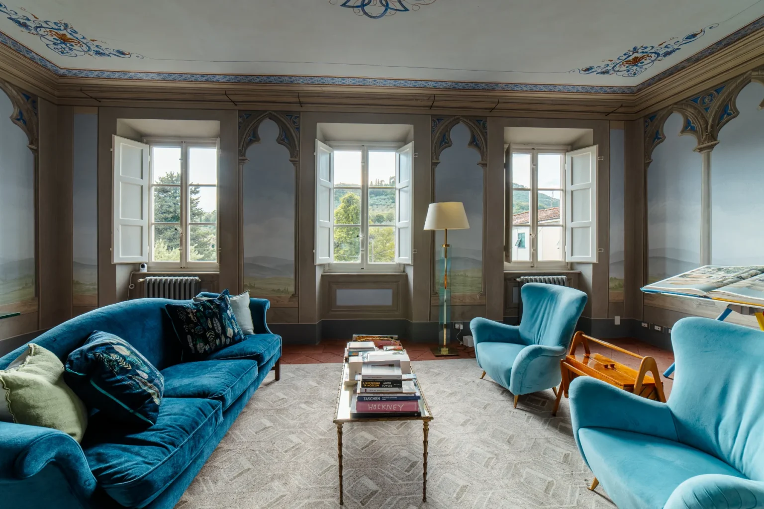 Elegant living room featuring a plush blue velvet sofa and armchairs, adorned with decorative pillows. The space is illuminated by natural light streaming through open windows, showcasing scenic views. A stylish coffee table displays a collection of art books atop a patterned area rug, with intricate wall murals and a modern floor lamp enhancing the ambiance. | Only on SopranoVillas