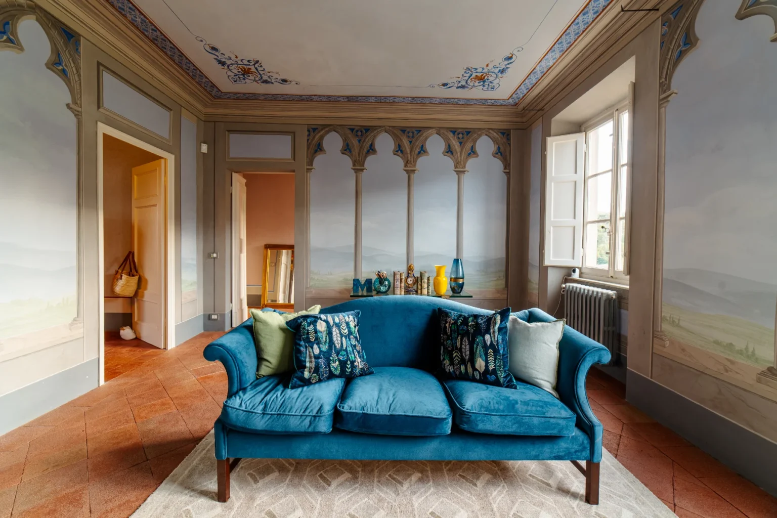Bright and inviting living room featuring a plush blue sofa adorned with decorative pillows, surrounded by elegant frescoed walls and large windows that provide natural light. The space includes a cozy area rug and decorative elements like vases and books, showcasing a blend of classic and modern design aesthetics. | Only on SopranoVillas