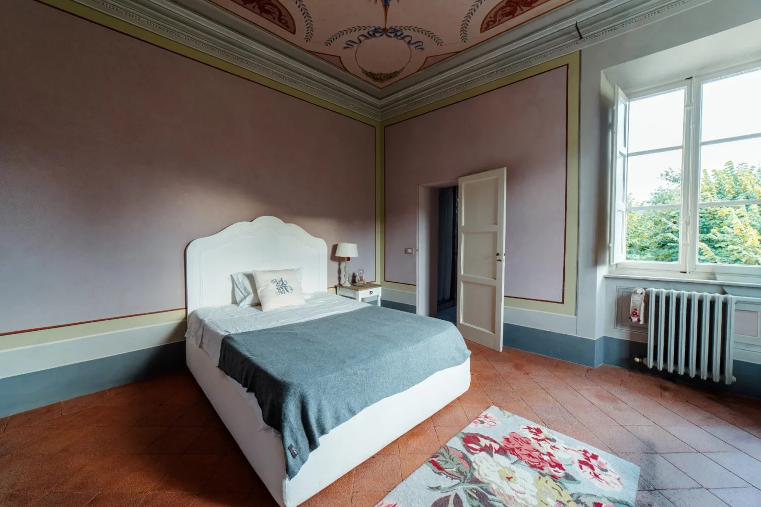 Spacious bedroom featuring a white bed with a gray throw, elegant ceiling details, and large windows allowing natural light. Decorated with a floral rug and a bedside table with a lamp, this room offers a serene and inviting atmosphere. | Only on SopranoVillas