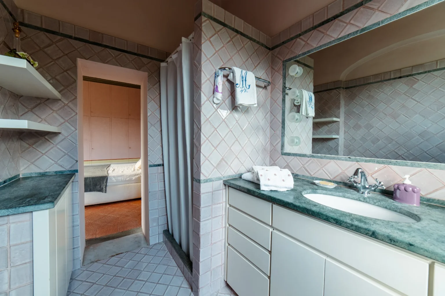 Modern bathroom interior featuring a green marble countertop, light pink tiled walls, and a shower area. The space includes a mirror, white cabinetry, and towels neatly arranged, with a view into an adjacent bedroom. | Only on SopranoVillas