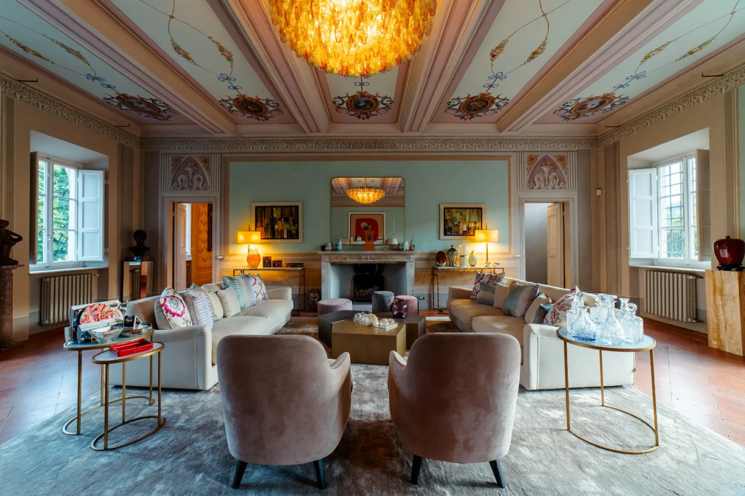 Luxurious living room featuring elegant sofas with decorative cushions, stylish armchairs, and a stunning chandelier. The room is adorned with intricate ceiling designs, framed artwork, and a marble fireplace, complemented by large windows allowing natural light to enhance the sophisticated ambiance. | Only on SopranoVillas
