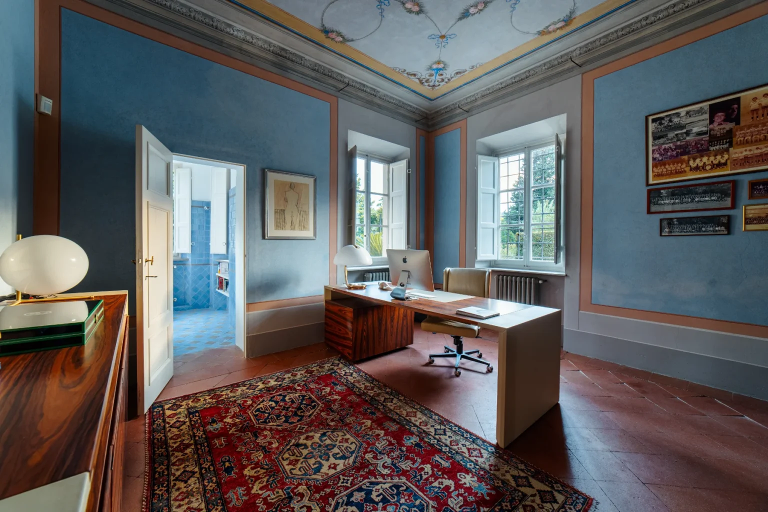 Bright and stylish home office featuring a modern desk with an iMac, surrounded by light blue walls with elegant molding. The room includes a decorative rug, a sleek lamp, and framed artwork, with large windows allowing natural light and a view of greenery outside. A doorway leads to a blue-tiled bathroom, enhancing the room's spacious feel. | Only on SopranoVillas