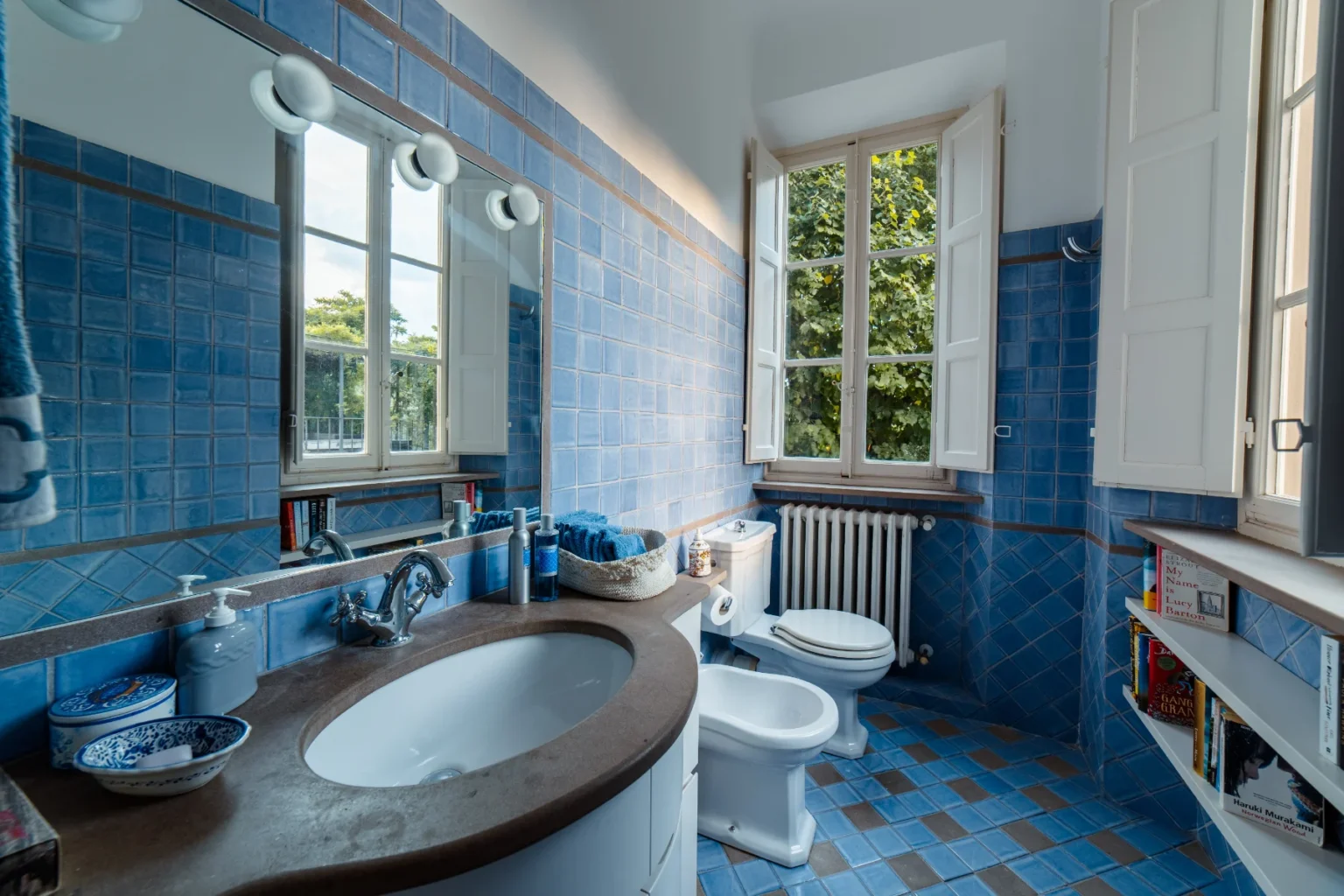Bright and airy bathroom featuring blue tiled walls and floor, a round sink with a stylish faucet, and a window with open shutters allowing natural light. The space includes a white toilet, bidet, and a shelf with books, creating a cozy and inviting atmosphere. | Only on SopranoVillas