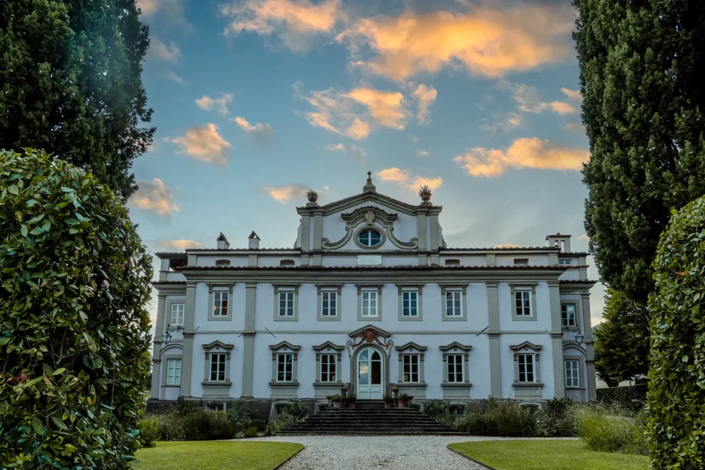 Historic villa with ornate architecture surrounded by lush greenery and a clear blue sky with clouds, showcasing a grand entrance and well-maintained gardens. | Only on SopranoVillas