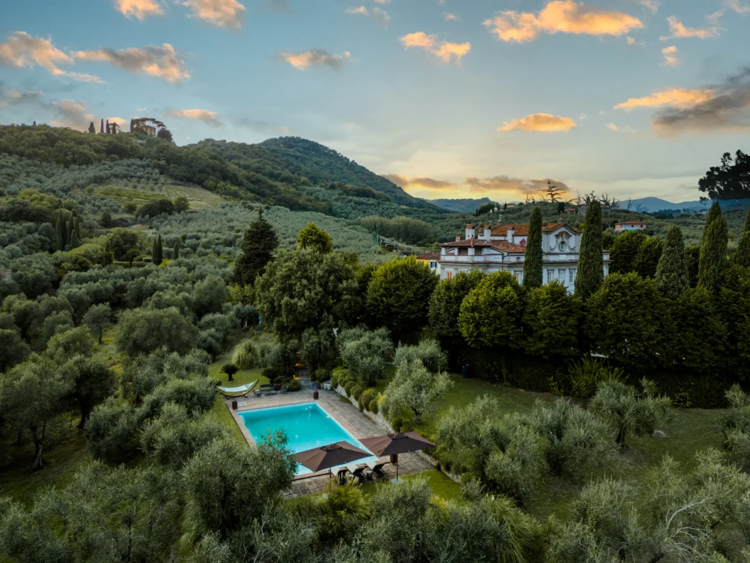 Aerial view of a serene villa surrounded by lush olive groves, featuring a sparkling swimming pool and landscaped gardens under a vibrant sunset sky. | Only on SopranoVillas