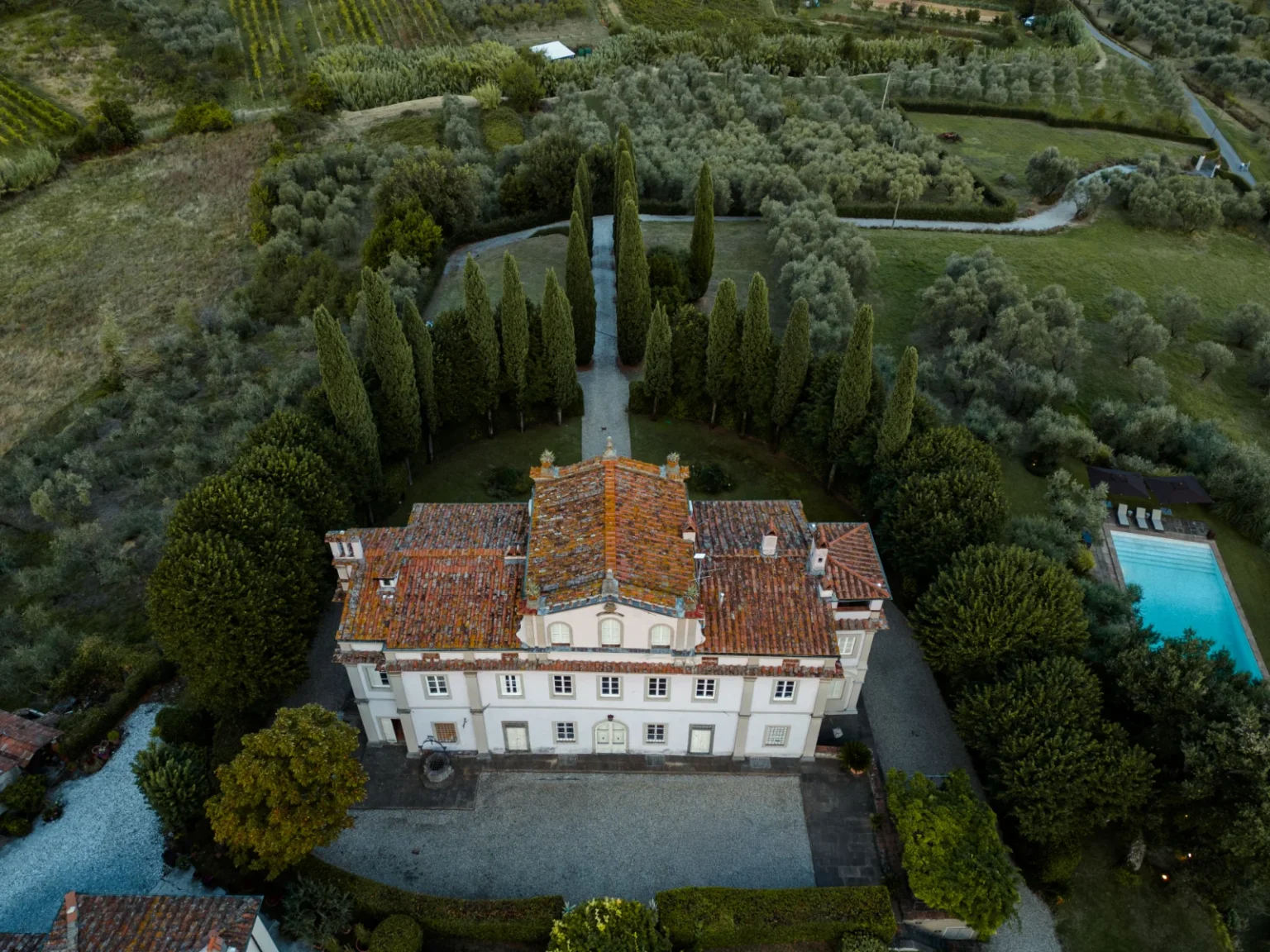 Aerial view of a charming villa surrounded by lush greenery, tall cypress trees, and a swimming pool, set in a picturesque landscape of vineyards and olive groves. | Only on SopranoVillas