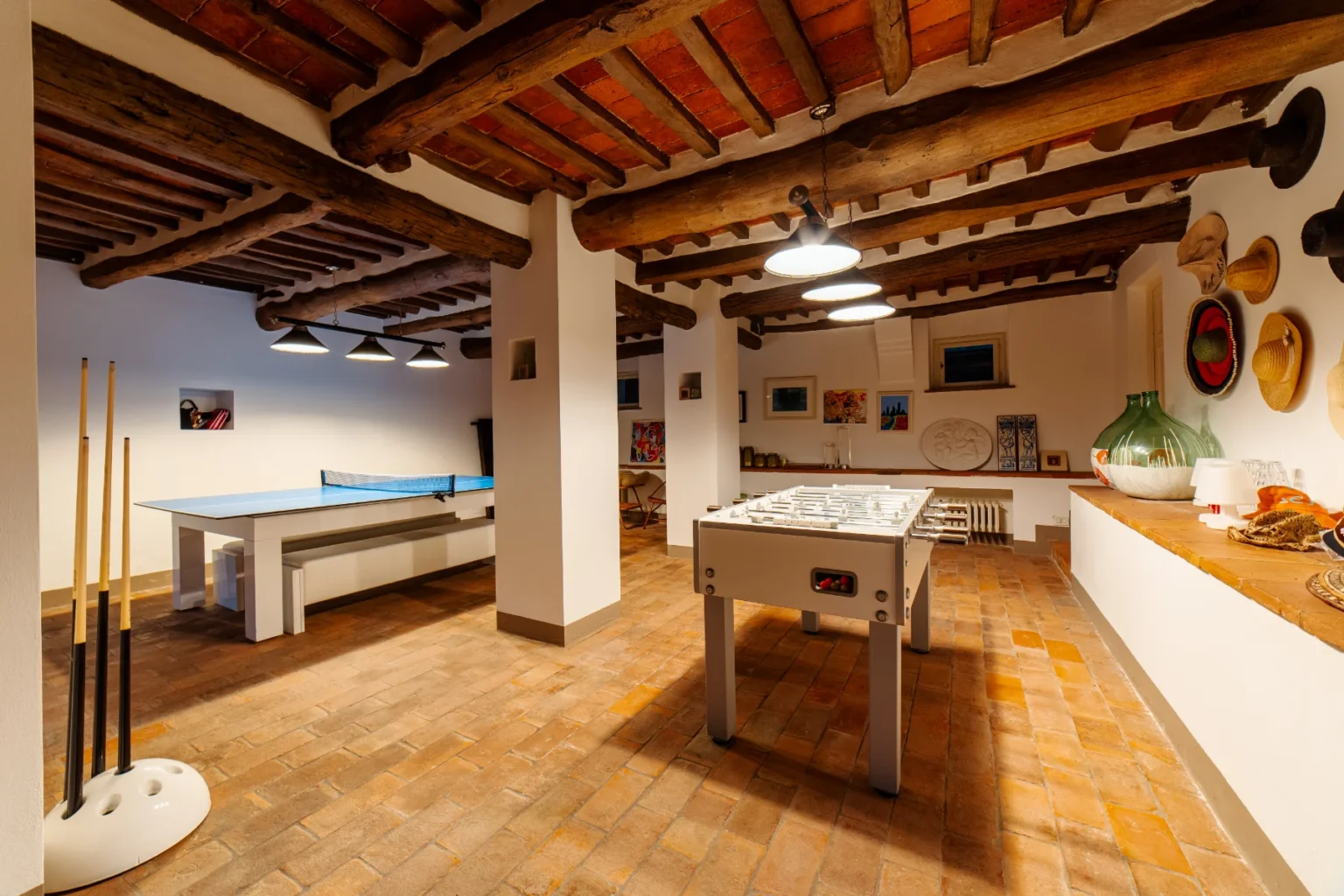 Spacious game room featuring a foosball table and a blue ping pong table, illuminated by modern pendant lights, with rustic wooden beams and terracotta tile flooring. Decorated with artwork and wall-mounted hats, creating a cozy and inviting atmosphere. | Only on SopranoVillas