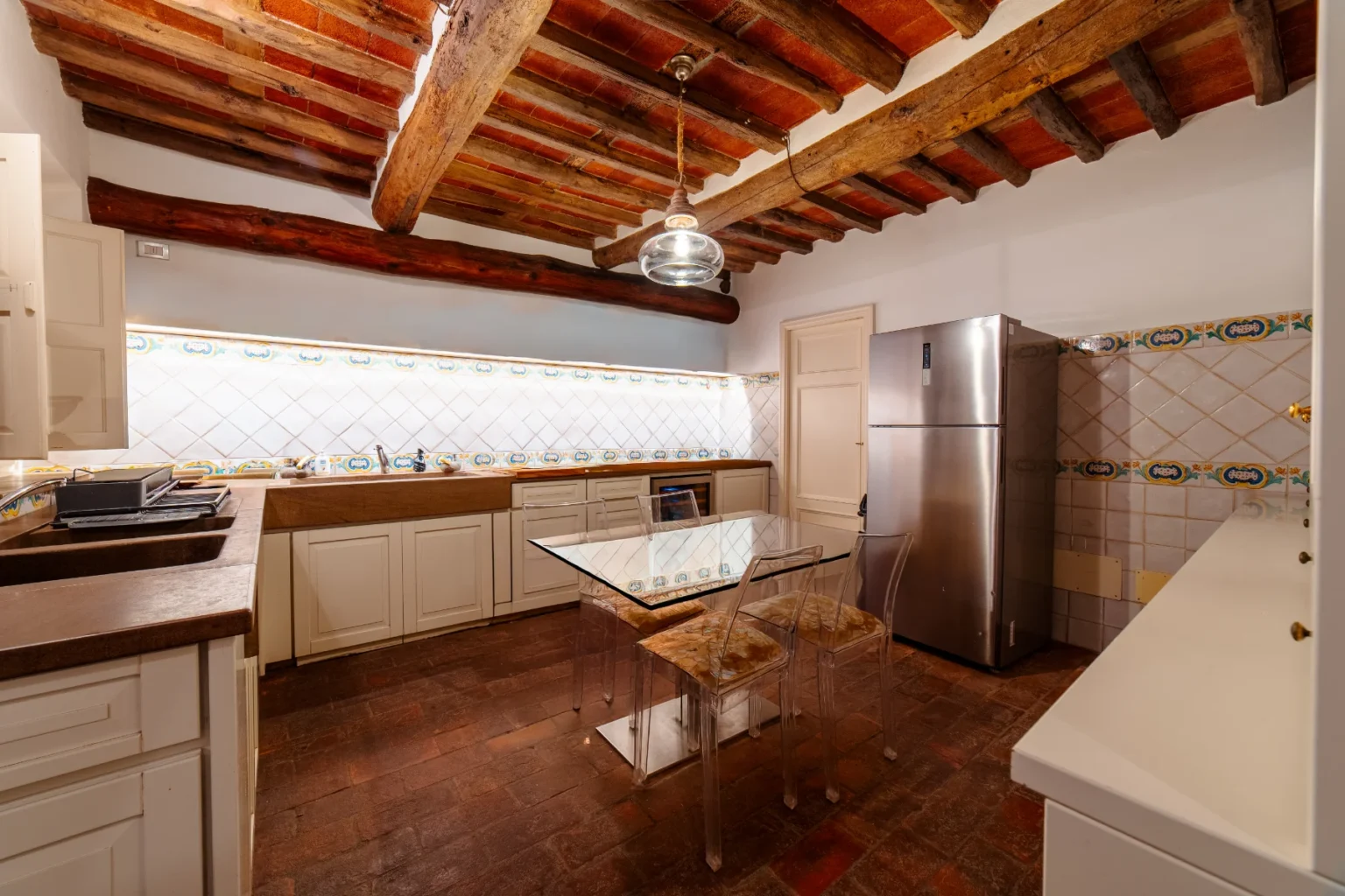 Modern kitchen with rustic wooden beams, featuring a glass dining table, clear acrylic chairs, a stainless steel refrigerator, and tiled backsplash. The warm terracotta floor complements the contemporary cabinetry and lighting. Ideal for cooking and entertaining. | Only on SopranoVillas