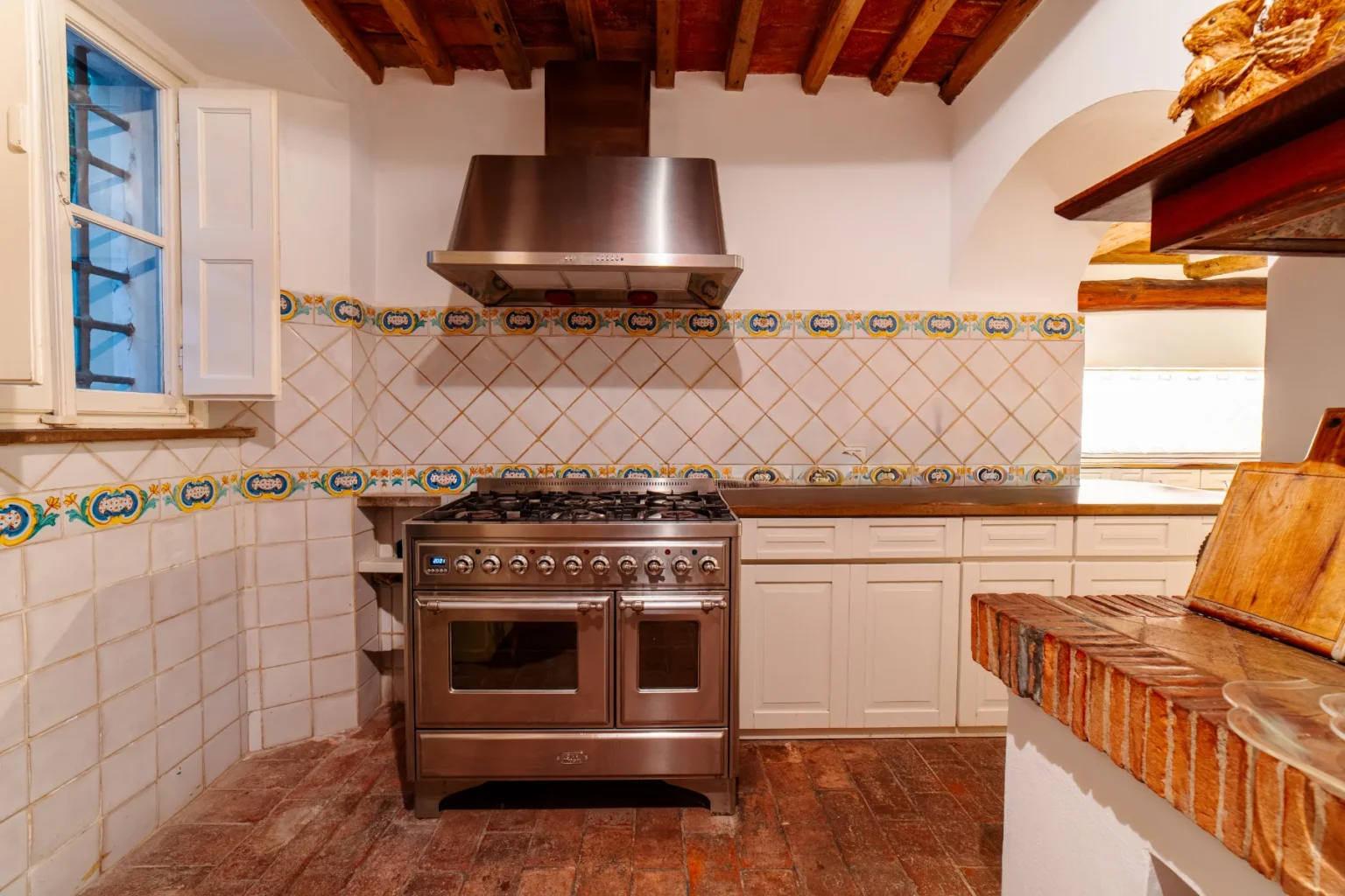 Rustic kitchen interior featuring a stainless steel gas range, a decorative tiled backsplash with floral patterns, and wooden ceiling beams. The space includes white cabinetry and a window that allows natural light to brighten the room. | Only on SopranoVillas
