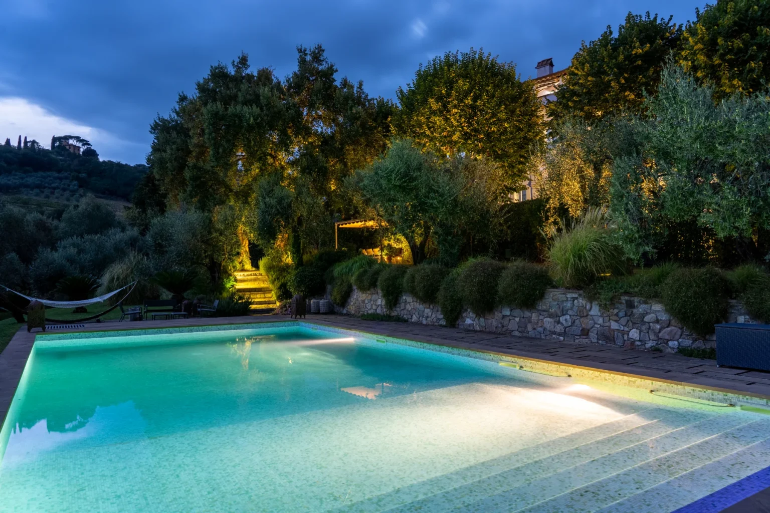 Serene nighttime view of a beautifully lit swimming pool surrounded by lush greenery and trees, with steps leading to a cozy outdoor seating area, creating a tranquil atmosphere perfect for relaxation. | Only on SopranoVillas