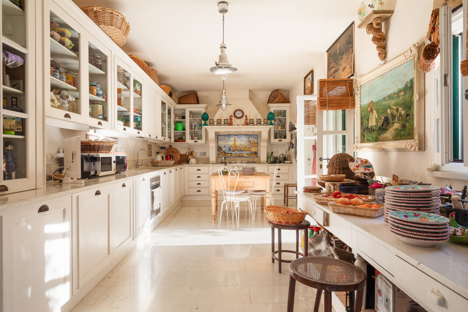 Bright and airy kitchen featuring white cabinetry, a wooden dining table, and various decorative baskets. The space includes modern appliances, colorful dishware, and a scenic painting on the wall, creating a warm and inviting atmosphere. Sunlight streams in, highlighting the clean design and organized storage. | Only on SopranoVillas