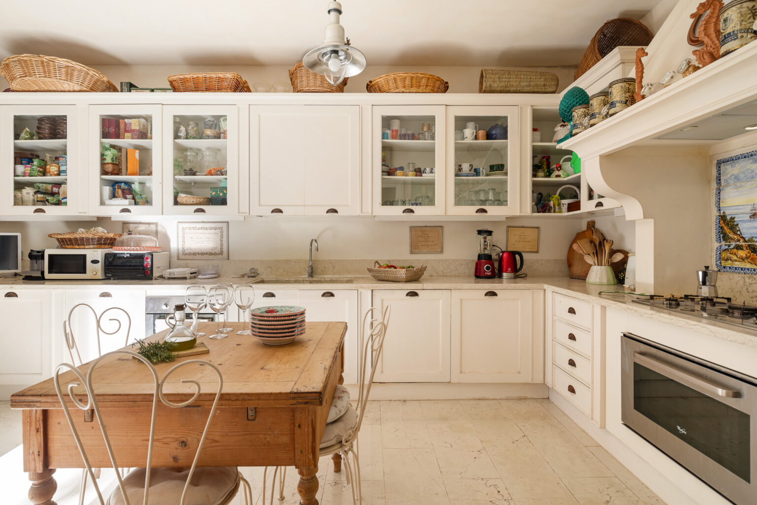 Bright and airy kitchen featuring white cabinetry, a wooden dining table, and various kitchen appliances. Shelves are filled with glassware and pantry items, while decorative baskets are placed on top. The space is well-organized, showcasing a blend of functionality and style. | Only on SopranoVillas