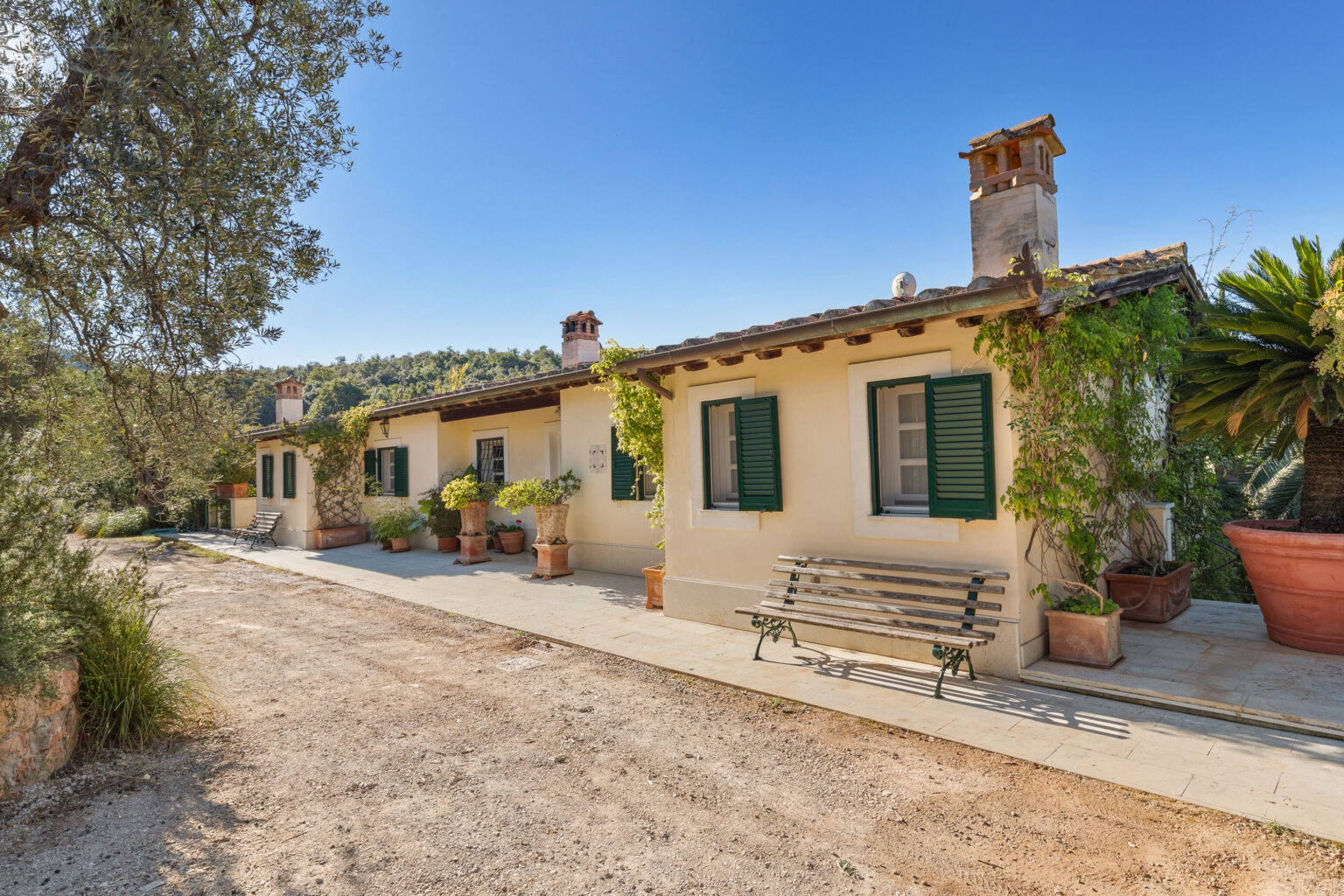 Charming rustic cottage with green shutters and a stone pathway, surrounded by potted plants and trees, set against a clear blue sky and rolling hills. Ideal for a serene getaway or vacation rental. | Only on SopranoVillas